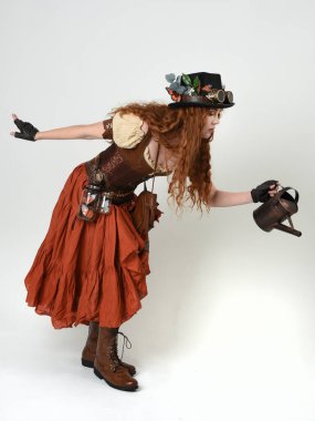 Uzun, yürüyen Steampunk Botanikçisi, çılgın bilim adamı, silindir şapkalı ve gözlüklü kadın model. Kızıl Saçlı Fantezi Karakteri. Elinde su dolu bir kutu var. İzole edilmiş Beyaz Stüdyo Arkaplanı 'nda silah gibi.