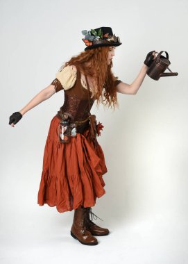 Uzun, yürüyen Steampunk Botanikçisi, çılgın bilim adamı, silindir şapkalı ve gözlüklü kadın model. Kızıl Saçlı Fantezi Karakteri. Elinde su dolu bir kutu var. İzole edilmiş Beyaz Stüdyo Arkaplanı 'nda silah gibi.