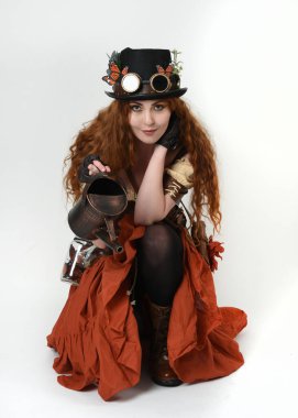 Uzun, yürüyen Steampunk Botanikçisi, çılgın bilim adamı, silindir şapkalı ve gözlüklü kadın model. Kızıl Saçlı Fantezi Karakteri. Elinde su dolu bir kutu var. İzole edilmiş Beyaz Stüdyo Arkaplanı 'nda silah gibi.