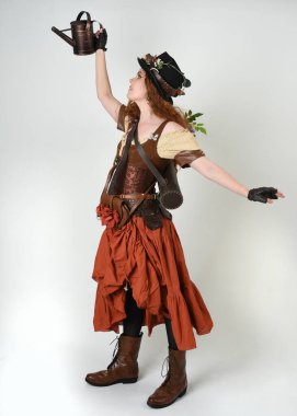 Uzun, yürüyen Steampunk Botanikçisi, çılgın bilim adamı, silindir şapkalı ve gözlüklü kadın model. Kızıl Saçlı Fantezi Karakteri. Elinde su dolu bir kutu var. İzole edilmiş Beyaz Stüdyo Arkaplanı 'nda silah gibi.