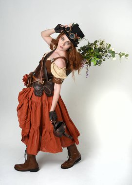 Uzun, yürüyen Steampunk Botanikçisi, çılgın bilim adamı, silindir şapkalı ve gözlüklü kadın model. Kızıl Saçlı Fantezi Karakteri. Elinde su dolu bir kutu var. İzole edilmiş Beyaz Stüdyo Arkaplanı 'nda silah gibi.