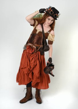 Uzun, yürüyen Steampunk Botanikçisi, çılgın bilim adamı, silindir şapkalı ve gözlüklü kadın model. Kızıl Saçlı Fantezi Karakteri. Elinde su dolu bir kutu var. İzole edilmiş Beyaz Stüdyo Arkaplanı 'nda silah gibi.