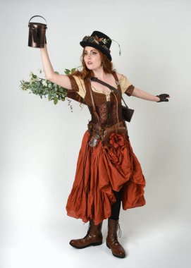 Uzun, yürüyen Steampunk Botanikçisi, çılgın bilim adamı, silindir şapkalı ve gözlüklü kadın model. Kızıl Saçlı Fantezi Karakteri. Elinde su dolu bir kutu var. İzole edilmiş Beyaz Stüdyo Arkaplanı 'nda silah gibi.