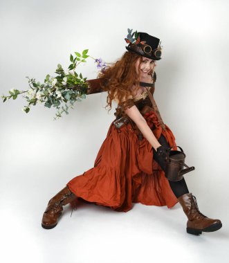 Uzun, yürüyen Steampunk Botanikçisi, çılgın bilim adamı, silindir şapkalı ve gözlüklü kadın model. Kızıl Saçlı Fantezi Karakteri. Elinde su dolu bir kutu var. İzole edilmiş Beyaz Stüdyo Arkaplanı 'nda silah gibi.