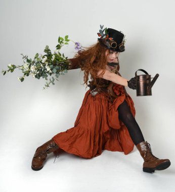 Uzun, yürüyen Steampunk Botanikçisi, çılgın bilim adamı, silindir şapkalı ve gözlüklü kadın model. Kızıl Saçlı Fantezi Karakteri. Elinde su dolu bir kutu var. İzole edilmiş Beyaz Stüdyo Arkaplanı 'nda silah gibi.