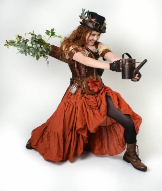 Uzun, yürüyen Steampunk Botanikçisi, çılgın bilim adamı, silindir şapkalı ve gözlüklü kadın model. Kızıl Saçlı Fantezi Karakteri. Elinde su dolu bir kutu var. İzole edilmiş Beyaz Stüdyo Arkaplanı 'nda silah gibi.