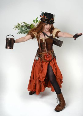 Uzun, yürüyen Steampunk Botanikçisi, çılgın bilim adamı, silindir şapkalı ve gözlüklü kadın model. Kızıl Saçlı Fantezi Karakteri. Elinde su dolu bir kutu var. İzole edilmiş Beyaz Stüdyo Arkaplanı 'nda silah gibi.