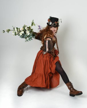 Uzun, yürüyen Steampunk Botanikçisi, çılgın bilim adamı, silindir şapkalı ve gözlüklü kadın model. Kızıl Saçlı Fantezi Karakteri. Elinde su dolu bir kutu var. İzole edilmiş Beyaz Stüdyo Arkaplanı 'nda silah gibi.