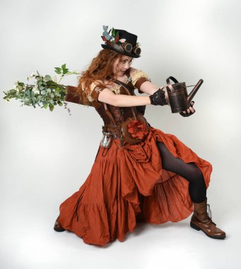 Uzun, yürüyen Steampunk Botanikçisi, çılgın bilim adamı, silindir şapkalı ve gözlüklü kadın model. Kızıl Saçlı Fantezi Karakteri. Elinde su dolu bir kutu var. İzole edilmiş Beyaz Stüdyo Arkaplanı 'nda silah gibi.