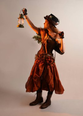 Uzun, yürüyen Steampunk Botanikçisi, çılgın bilim adamı, silindir şapkalı ve gözlüklü kadın model. Kızıl Saçlı Fantezi Karakteri. Elinde bir fener, stüdyo arka planı var.