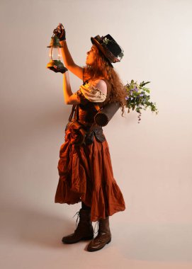 Uzun, yürüyen Steampunk Botanikçisi, çılgın bilim adamı, silindir şapkalı ve gözlüklü kadın model. Kızıl Saçlı Fantezi Karakteri. Elinde bir fener, stüdyo arka planı var.