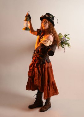 Uzun, yürüyen Steampunk Botanikçisi, çılgın bilim adamı, silindir şapkalı ve gözlüklü kadın model. Kızıl Saçlı Fantezi Karakteri. Elinde bir fener, stüdyo arka planı var.