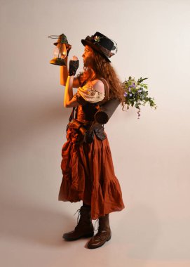 Uzun, yürüyen Steampunk Botanikçisi, çılgın bilim adamı, silindir şapkalı ve gözlüklü kadın model. Kızıl Saçlı Fantezi Karakteri. Elinde bir fener, stüdyo arka planı var.