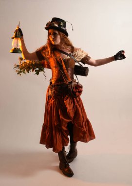 Uzun, yürüyen Steampunk Botanikçisi, çılgın bilim adamı, silindir şapkalı ve gözlüklü kadın model. Kızıl Saçlı Fantezi Karakteri. Elinde bir fener, stüdyo arka planı var.