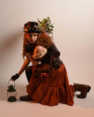 Uzun, yürüyen Steampunk Botanikçisi, çılgın bilim adamı, silindir şapkalı ve gözlüklü kadın model. Kızıl Saçlı Fantezi Karakteri. Elinde bir fener, stüdyo arka planı var.