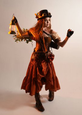 Uzun, yürüyen Steampunk Botanikçisi, çılgın bilim adamı, silindir şapkalı ve gözlüklü kadın model. Kızıl Saçlı Fantezi Karakteri. Elinde bir fener, stüdyo arka planı var.