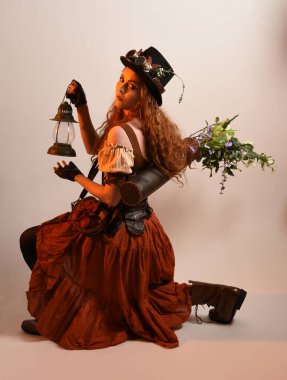 Uzun, yürüyen Steampunk Botanikçisi, çılgın bilim adamı, silindir şapkalı ve gözlüklü kadın model. Kızıl Saçlı Fantezi Karakteri. Elinde bir fener, stüdyo arka planı var.