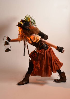 Uzun, yürüyen Steampunk Botanikçisi, çılgın bilim adamı, silindir şapkalı ve gözlüklü kadın model. Kızıl Saçlı Fantezi Karakteri. Elinde bir fener, stüdyo arka planı var.