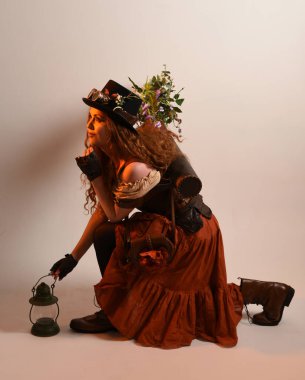 Uzun, yürüyen Steampunk Botanikçisi, çılgın bilim adamı, silindir şapkalı ve gözlüklü kadın model. Kızıl Saçlı Fantezi Karakteri. Elinde bir fener, stüdyo arka planı var.