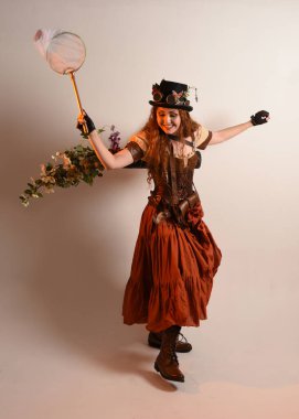 Uzun, yürüyen Steampunk Botanikçisi, çılgın bilim adamı, silindir şapkalı ve gözlüklü kadın model. Kızıl Saçlı Fantezi Karakteri Poz Kelebek Avlama Ağı, Stüdyo Arkaplan