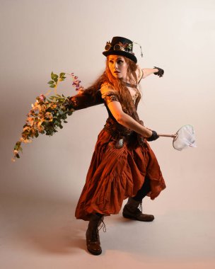 Uzun, yürüyen Steampunk Botanikçisi, çılgın bilim adamı, silindir şapkalı ve gözlüklü kadın model. Kızıl Saçlı Fantezi Karakteri Poz Kelebek Avlama Ağı, Stüdyo Arkaplan