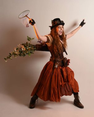 Uzun, yürüyen Steampunk Botanikçisi, çılgın bilim adamı, silindir şapkalı ve gözlüklü kadın model. Kızıl Saçlı Fantezi Karakteri Poz Kelebek Avlama Ağı, Stüdyo Arkaplan