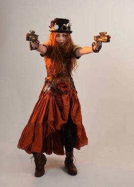 Uzun, yürüyen Steampunk Botanikçisi, çılgın bilim adamı, silindir şapkalı ve gözlüklü kadın model. Kızıl Saçlı Fantezi Karakteri. Elinde silah olan bir poz, stüdyo arka planı.