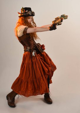 Uzun, yürüyen Steampunk Botanikçisi, çılgın bilim adamı, silindir şapkalı ve gözlüklü kadın model. Kızıl Saçlı Fantezi Karakteri. Elinde silah olan bir poz, stüdyo arka planı.