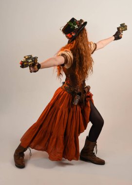 Uzun, yürüyen Steampunk Botanikçisi, çılgın bilim adamı, silindir şapkalı ve gözlüklü kadın model. Kızıl Saçlı Fantezi Karakteri. Elinde silah olan bir poz, stüdyo arka planı.