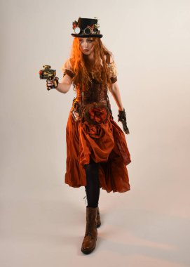 Uzun, yürüyen Steampunk Botanikçisi, çılgın bilim adamı, silindir şapkalı ve gözlüklü kadın model. Kızıl Saçlı Fantezi Karakteri. Elinde silah olan bir poz, stüdyo arka planı.