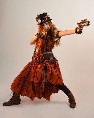 Uzun, yürüyen Steampunk Botanikçisi, çılgın bilim adamı, silindir şapkalı ve gözlüklü kadın model. Kızıl Saçlı Fantezi Karakteri. Elinde silah olan bir poz, stüdyo arka planı.