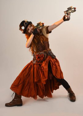 Uzun, yürüyen Steampunk Botanikçisi, çılgın bilim adamı, silindir şapkalı ve gözlüklü kadın model. Kızıl Saçlı Fantezi Karakteri. Elinde silah olan bir poz, stüdyo arka planı.