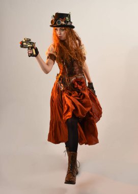 Uzun, yürüyen Steampunk Botanikçisi, çılgın bilim adamı, silindir şapkalı ve gözlüklü kadın model. Kızıl Saçlı Fantezi Karakteri. Elinde silah olan bir poz, stüdyo arka planı.
