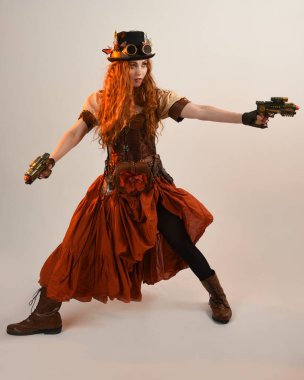 Uzun, yürüyen Steampunk Botanikçisi, çılgın bilim adamı, silindir şapkalı ve gözlüklü kadın model. Kızıl Saçlı Fantezi Karakteri. Elinde silah olan bir poz, stüdyo arka planı.