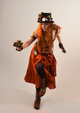 Uzun, yürüyen Steampunk Botanikçisi, çılgın bilim adamı, silindir şapkalı ve gözlüklü kadın model. Kızıl Saçlı Fantezi Karakteri. Elinde silah olan bir poz, stüdyo arka planı.