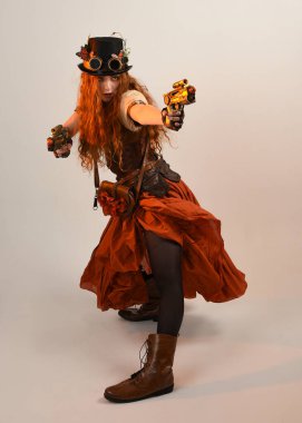 Uzun, yürüyen Steampunk Botanikçisi, çılgın bilim adamı, silindir şapkalı ve gözlüklü kadın model. Kızıl Saçlı Fantezi Karakteri. Elinde silah olan bir poz, stüdyo arka planı.