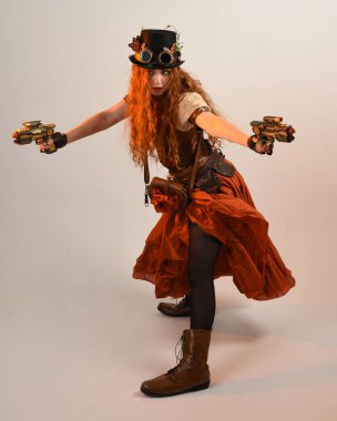 Uzun, yürüyen Steampunk Botanikçisi, çılgın bilim adamı, silindir şapkalı ve gözlüklü kadın model. Kızıl Saçlı Fantezi Karakteri. Elinde silah olan bir poz, stüdyo arka planı.