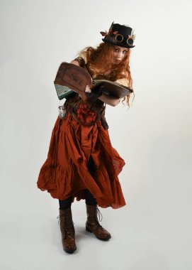 Uzun, yürüyen Steampunk Botanikçisi, çılgın bilim adamı, silindir şapkalı ve gözlüklü kadın model. Kızıl Saçlı Fantezi Karakteri Poz Beyaz Stüdyo Arkaplanı hakkında akademik kitaplar tutuyor