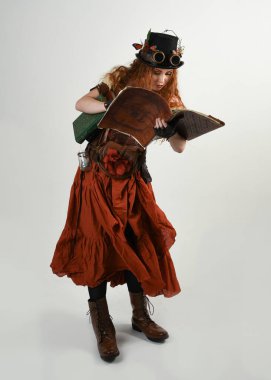 Uzun, yürüyen Steampunk Botanikçisi, çılgın bilim adamı, silindir şapkalı ve gözlüklü kadın model. Kızıl Saçlı Fantezi Karakteri Poz Beyaz Stüdyo Arkaplanı hakkında akademik kitaplar tutuyor
