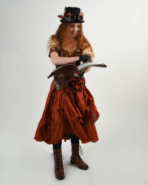 Uzun, yürüyen Steampunk Botanikçisi, çılgın bilim adamı, silindir şapkalı ve gözlüklü kadın model. Kızıl Saçlı Fantezi Karakteri Poz Beyaz Stüdyo Arkaplanı hakkında akademik kitaplar tutuyor