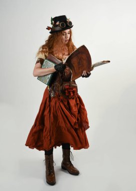 Uzun, yürüyen Steampunk Botanikçisi, çılgın bilim adamı, silindir şapkalı ve gözlüklü kadın model. Kızıl Saçlı Fantezi Karakteri Poz Beyaz Stüdyo Arkaplanı hakkında akademik kitaplar tutuyor