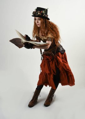 Uzun, yürüyen Steampunk Botanikçisi, çılgın bilim adamı, silindir şapkalı ve gözlüklü kadın model. Kızıl Saçlı Fantezi Karakteri Poz Beyaz Stüdyo Arkaplanı hakkında akademik kitaplar tutuyor