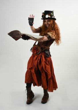 Uzun, yürüyen Steampunk Botanikçisi, çılgın bilim adamı, silindir şapkalı ve gözlüklü kadın model. Kızıl Saçlı Fantezi Karakteri Poz Beyaz Stüdyo Arkaplanı hakkında akademik kitaplar tutuyor