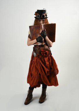 Uzun, yürüyen Steampunk Botanikçisi, çılgın bilim adamı, silindir şapkalı ve gözlüklü kadın model. Kızıl Saçlı Fantezi Karakteri Poz Beyaz Stüdyo Arkaplanı hakkında akademik kitaplar tutuyor