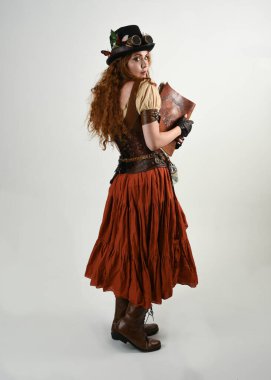 Uzun, yürüyen Steampunk Botanikçisi, çılgın bilim adamı, silindir şapkalı ve gözlüklü kadın model. Kızıl Saçlı Fantezi Karakteri Poz Beyaz Stüdyo Arkaplanı hakkında akademik kitaplar tutuyor