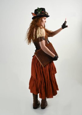 Uzun, yürüyen Steampunk Botanikçisi, çılgın bilim adamı, silindir şapkalı ve gözlüklü kadın model. Kızıl Saçlı Fantezi Karakteri Poz Beyaz Stüdyo Arkaplanı hakkında akademik kitaplar tutuyor