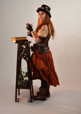 Uzun, yürüyen Steampunk Botanikçisi, çılgın bilim adamı, silindir şapkalı ve gözlüklü kadın model. Kızıl Saçlı Fantezi Karakteri Poz Beyaz Stüdyo Arkaplanı hakkında akademik kitaplar tutuyor