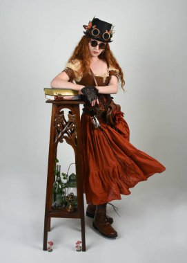 Uzun, yürüyen Steampunk Botanikçisi, çılgın bilim adamı, silindir şapkalı ve gözlüklü kadın model. Kızıl Saçlı Fantezi Karakteri Poz Beyaz Stüdyo Arkaplanı hakkında akademik kitaplar tutuyor