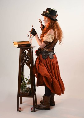 Uzun, yürüyen Steampunk Botanikçisi, çılgın bilim adamı, silindir şapkalı ve gözlüklü kadın model. Kızıl Saçlı Fantezi Karakteri Poz Beyaz Stüdyo Arkaplanı hakkında akademik kitaplar tutuyor