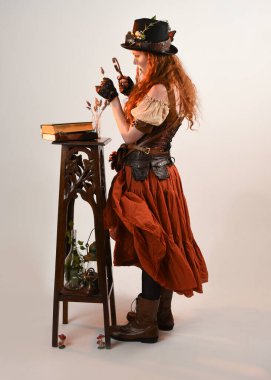Uzun, yürüyen Steampunk Botanikçisi, çılgın bilim adamı, silindir şapkalı ve gözlüklü kadın model. Kızıl Saçlı Fantezi Karakteri Poz Beyaz Stüdyo Arkaplanı hakkında akademik kitaplar tutuyor